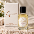 A bottle of Agare Ruby Eau de Parfum with a black cap stands next to its cream-colored packaging box on a polished stone slab, with soft white flowers blurred in the background.
